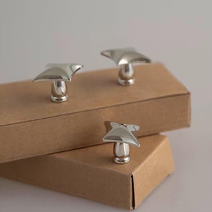 以下が含まれることがあります： 茶色の段ボール箱の上に、銀色の星型のキャビネットノブが3つあります。ノブは研磨仕上げで、モダンなデザインです。背景はニュートラルなオフホワイトです。
