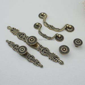 Könnte beinhalten: Eine Auswahl an antiken Messing-Schrankbeschlägen, darunter Griffe und Knöpfe. Die Beschläge weisen aufwendige Blumen- und Scrollarbeiten mit einem zentralen sonnenblumenartigen Motiv auf. Die Teile variieren in Größe und Form, geeignet für Schubladen und Türen.