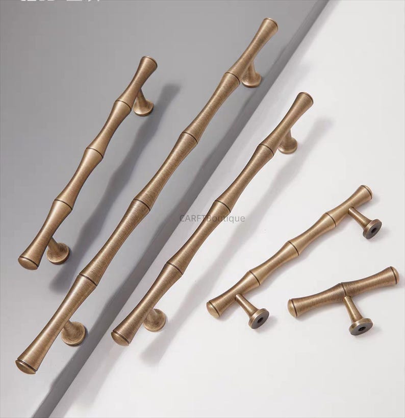 K&ouml;nnte beinhalten: Bronzefarbene Schrankgriffe im Bambus-Design. Die Griffe variieren in Gr&ouml;&szlig;e und Form und haben eine naturalistische &Auml;sthetik. Die Hardware ist auf einem wei&szlig;en und grauen Hintergrund angeordnet und zeigt die verschiedenen Gr&ouml;&szlig;en und Stile.