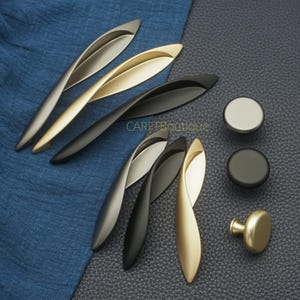May include: An assortment of cabinet hardware, including leaf-shaped pulls in silver, gold, and black finishes, along with round knobs in silver, black, and gold. The hardware is displayed on a blue cloth and a dark gray surface.