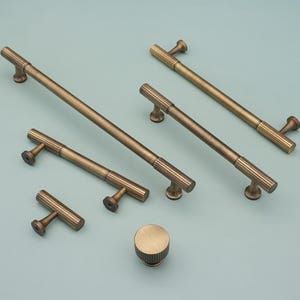 May include: An assortment of antique brass cabinet hardware, including several bar pulls and a round knob. The pulls vary in size, with a ribbed center section and rounded ends. The knob has a matching ribbed design.