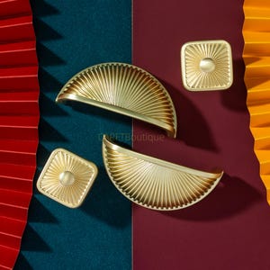 Puede incluir: Juego de herrajes dorados para armarios, que incluye dos tiradores semicirculares y dos pomos cuadrados. Los herrajes presentan un diseño de rayos de sol y se exhiben sobre un fondo de abanicos de papel turquesa, burdeos y naranja.