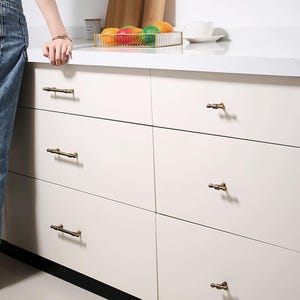 K&ouml;nnte beinhalten: Cremefarbene Schrankausz&uuml;ge mit antiker Messingbeschl&auml;gen. Die Schubladen sind in einem Raster angeordnet, wobei sechs sichtbar sind. Die Hardware ist ein Stangengriff. Die Arbeitsplatte ist wei&szlig;, und ein goldfarbener Drahtkorb mit Obst ist sichtbar.