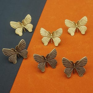 May include: Six decorative butterfly-shaped objects in varying shades of gold and bronze. The butterflies are arranged on a two-toned background of dark blue-gray and orange. Each butterfly has detailed wing patterns.