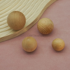 May include: Four wooden spheres of varying sizes are arranged on a pink surface, with a wooden tray in the background. The spheres have a natural wood grain pattern. The text "CARFTBoutique" is visible.