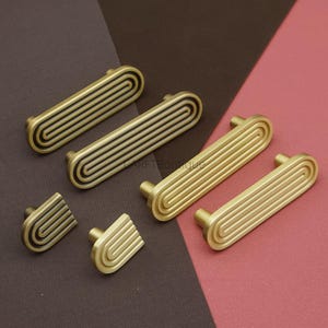 Puede incluir: Un conjunto de tiradores y pomos de armario en tono lat&oacute;n. Los tiradores son rectangulares con un dise&ntilde;o acanalado, y los pomos tienen un patr&oacute;n curvo y rayado. La ferreter&iacute;a se encuentra sobre un fondo marr&oacute;n y rosa.