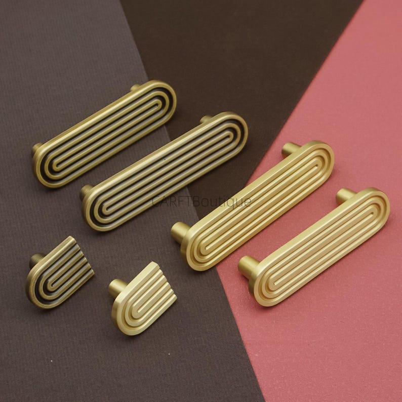 Puede incluir: Un conjunto de tiradores y pomos de armario en tono lat&oacute;n. Los tiradores son rectangulares con un dise&ntilde;o acanalado, y los pomos tienen un patr&oacute;n curvo y rayado. La ferreter&iacute;a se encuentra sobre un fondo marr&oacute;n y rosa.