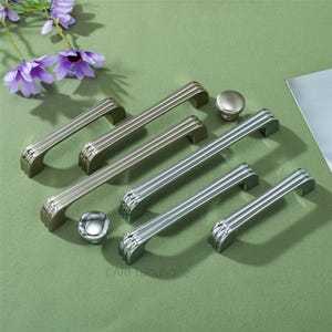 Puede incluir: Una colección de tiradores y pomos de armario plateados y bronceados. Los tiradores tienen un diseño acanalado y están dispuestos con dos pomos redondos. El hardware se muestra sobre una superficie verde con flores moradas en el fondo.