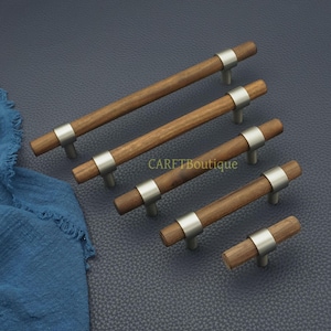 以下が含まれることがあります： 木製のキャビネットプルセット。つや消し金属のアクセントが付いています。各プルは、中央に銀色のバンドが付いた円筒形の木製ハンドルを備えています。「CARETBoutique」の文字が背景に見えます。