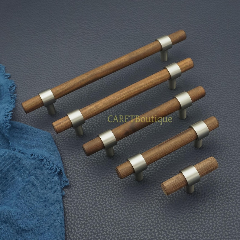 以下が含まれることがあります： 木製のキャビネットプルセット。つや消し金属のアクセントが付いています。各プルは、中央に銀色のバンドが付いた円筒形の木製ハンドルを備えています。「CARETBoutique」の文字が背景に見えます。