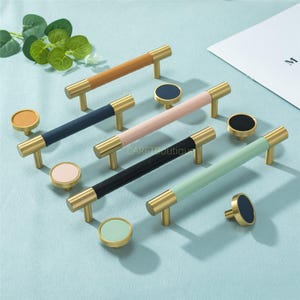 May include: A selection of cabinet handles and knobs. The handles have gold-coloured metal ends and leather-wrapped centres in tan, navy, pink, black, and mint green. Matching knobs are also included, all in a range of colours.