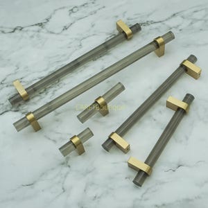 Peut inclure: Ensemble de poignées et de boutons de placard en acrylique gris transparent avec des accents métalliques dorés. Les poignées sont de différentes longueurs et les boutons sont cylindriques. Le matériel est disposé sur une surface à motifs de marbre.