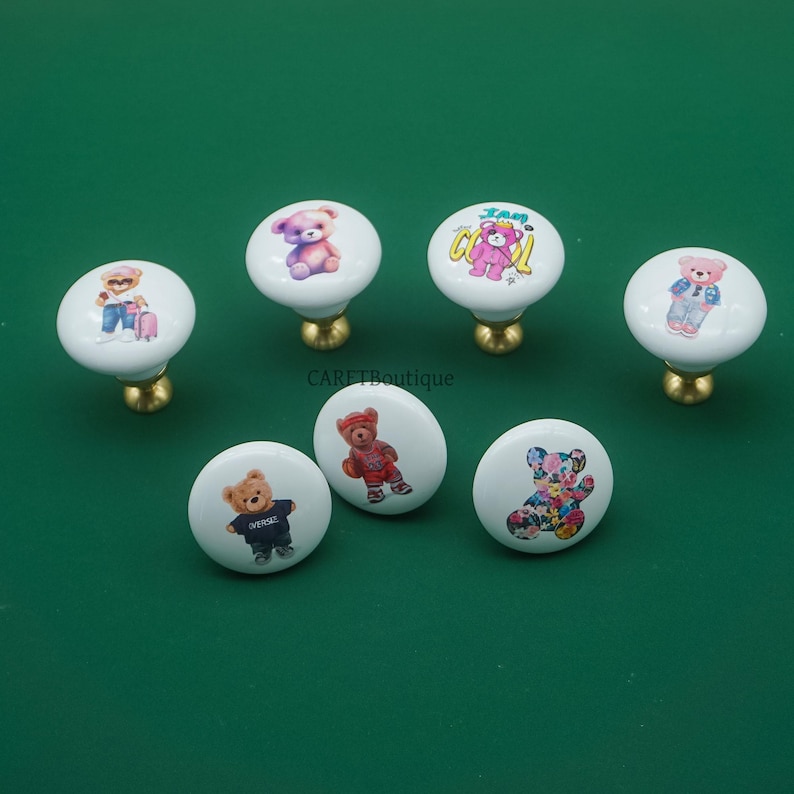 May include: Seven white ceramic cabinet knobs with colorful teddy bear designs. Each knob has a gold-colored base. The bears are depicted in various outfits and styles, including a bear with the text "I am cool".