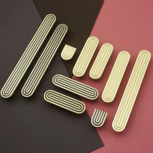 Puede incluir: Un conjunto de tiradores y pomos dorados para armarios con un dise&ntilde;o acanalado. Los tiradores son rectangulares, y los pomos son ovalados y en forma de U. Las ranuras son negras. El hardware est&aacute; dispuesto sobre un fondo burdeos y rosa.