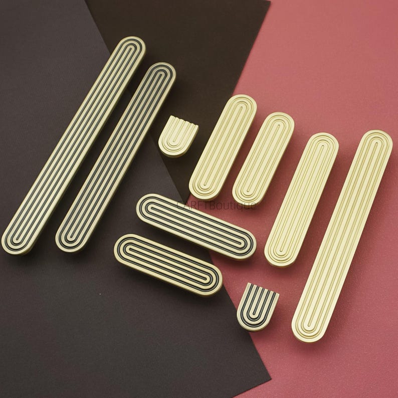 Puede incluir: Un conjunto de tiradores y pomos dorados para armarios con un dise&ntilde;o acanalado. Los tiradores son rectangulares, y los pomos son ovalados y en forma de U. Las ranuras son negras. El hardware est&aacute; dispuesto sobre un fondo burdeos y rosa.