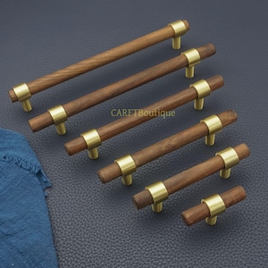 以下が含まれることがあります： 真鍮色のアクセントが付いた木製キャビネットプル。プルは円筒形の木製ハンドルで、真鍮色のリングと2つの小さな真鍮色のポストでキャビネットに接続されています。「CARFTBoutique」の文字が見えます。