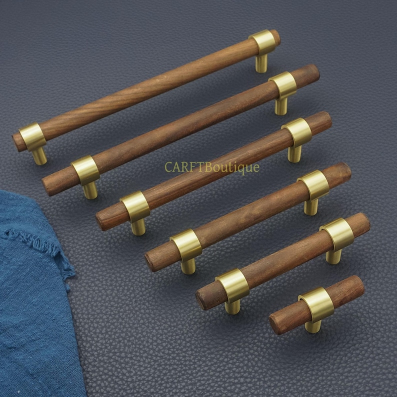 以下が含まれることがあります： 真鍮色のアクセントが付いた木製キャビネットプル。プルは円筒形の木製ハンドルで、真鍮色のリングと2つの小さな真鍮色のポストでキャビネットに接続されています。「CARFTBoutique」の文字が見えます。