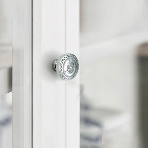 May include: A close-up of a silver cabinet knob with an intricate circular design. The knob is mounted on a white cabinet door. The design features geometric and floral patterns.