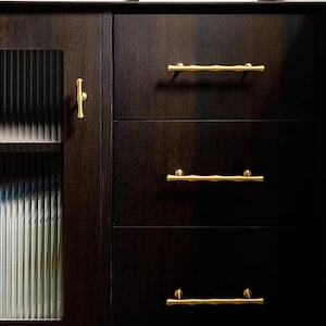 K&ouml;nnte beinhalten: Dunkler Holzschrank mit drei Schubladen und einer Glast&uuml;r. Der Schrank hat goldfarbene, astf&ouml;rmige Schubladengriffe und einen passenden Griff an der T&uuml;r. Das Holz hat eine satte, dunkelbraune Oberfl&auml;che.