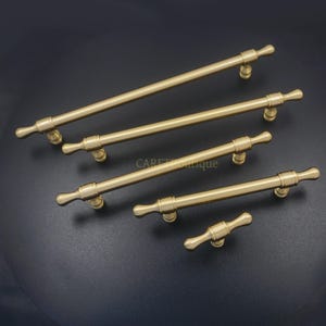 K&ouml;nnte beinhalten: F&uuml;nf messingfarbene Schrankgriffe in verschiedenen Gr&ouml;&szlig;en. Jeder Griff hat eine zylindrische Stange mit dekorativen Endst&uuml;cken und einer gebogenen Basis zur Montage. Die Griffe sind diagonal vor einem dunklen Hintergrund angeordnet und zeigen ihr Design und ihre Oberfl&auml;che.