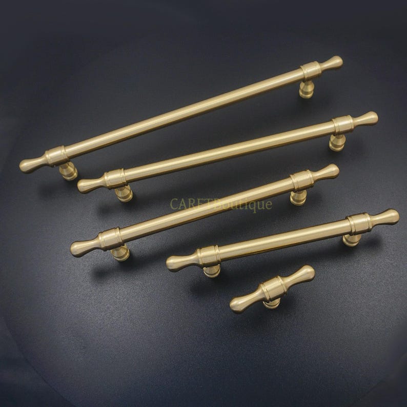 K&ouml;nnte beinhalten: F&uuml;nf messingfarbene Schrankgriffe in verschiedenen Gr&ouml;&szlig;en. Jeder Griff hat eine zylindrische Stange mit dekorativen Endst&uuml;cken und einer gebogenen Basis zur Montage. Die Griffe sind diagonal vor einem dunklen Hintergrund angeordnet und zeigen ihr Design und ihre Oberfl&auml;che.