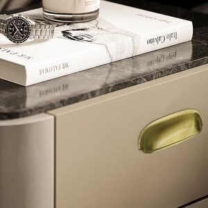 May include: A silver wristwatch with a black dial sits on a white book titled "Mr. Palomar" and "Italo Calvino". A candle sits atop a marble surface. A gold-toned drawer pull is visible.