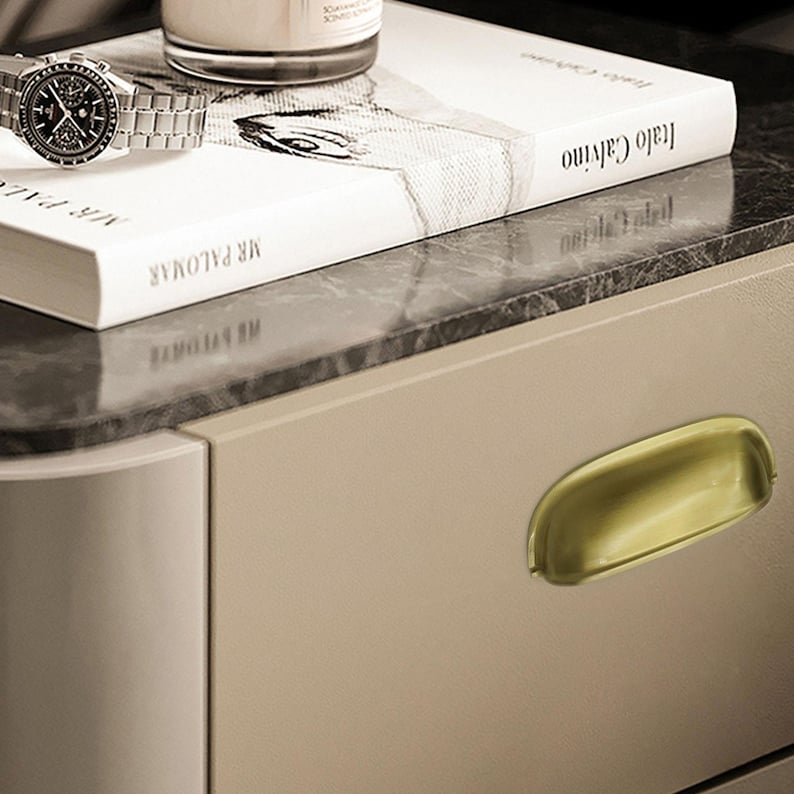 May include: A silver wristwatch with a black dial sits on a white book titled "Mr. Palomar" and "Italo Calvino". A candle sits atop a marble surface. A gold-toned drawer pull is visible.