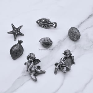 Peut inclure: Une collection de petits objets d&eacute;coratifs argent&eacute;s. L'assortiment comprend une &eacute;toile de mer, un poisson, un coquillage et un ch&eacute;rubin tenant un parchemin avec le mot "Luck". Les objets sont dispos&eacute;s sur une surface en marbre blanc.