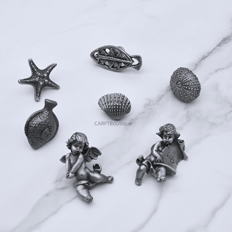 Peut inclure: Une collection de petits objets d&eacute;coratifs argent&eacute;s. L'assortiment comprend une &eacute;toile de mer, un poisson, un coquillage et un ch&eacute;rubin tenant un parchemin avec le mot "Luck". Les objets sont dispos&eacute;s sur une surface en marbre blanc.