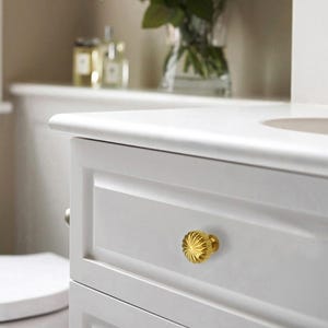May include: Close-up of a white cabinet with a gold-colored, fluted drawer knob. The knob has a detailed, sunburst design. The cabinet is part of a bathroom vanity set.