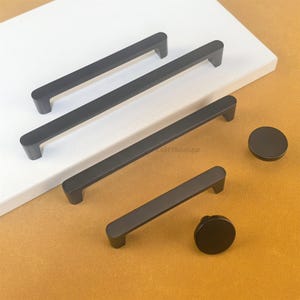 May include: A selection of matte black cabinet hardware, featuring four rectangular bar pulls and two round knobs. The pulls are of varying lengths, and the knobs are circular. The hardware is presented on a white surface against a tan backdrop.
