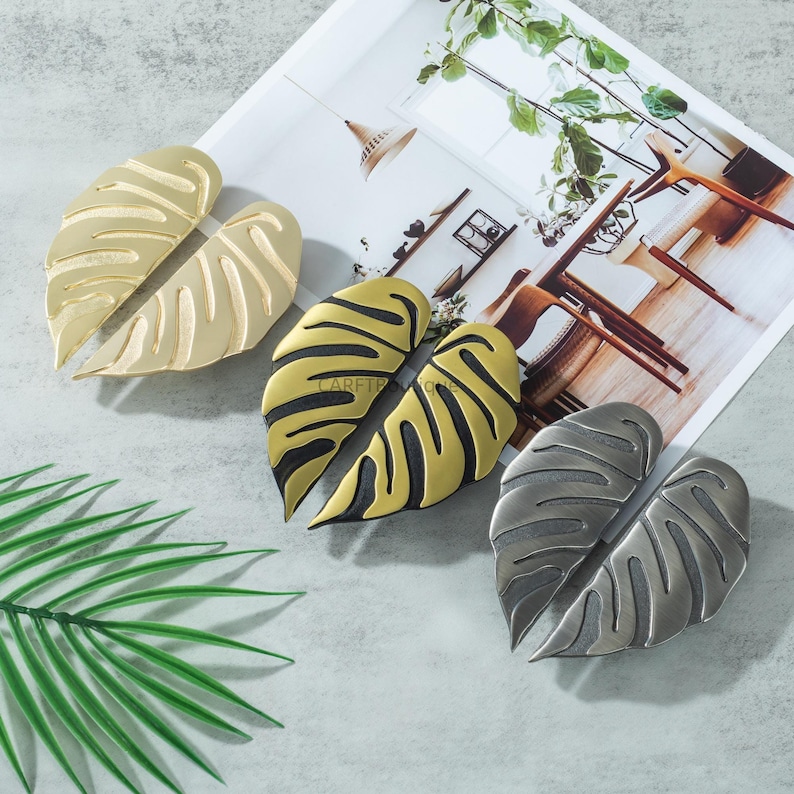 May include: Decorative leaf-shaped cabinet pulls in gold, black and gold, and silver finishes. The pulls are arranged on a light gray surface with a magazine and a green leaf in the background. The pulls have a textured design.