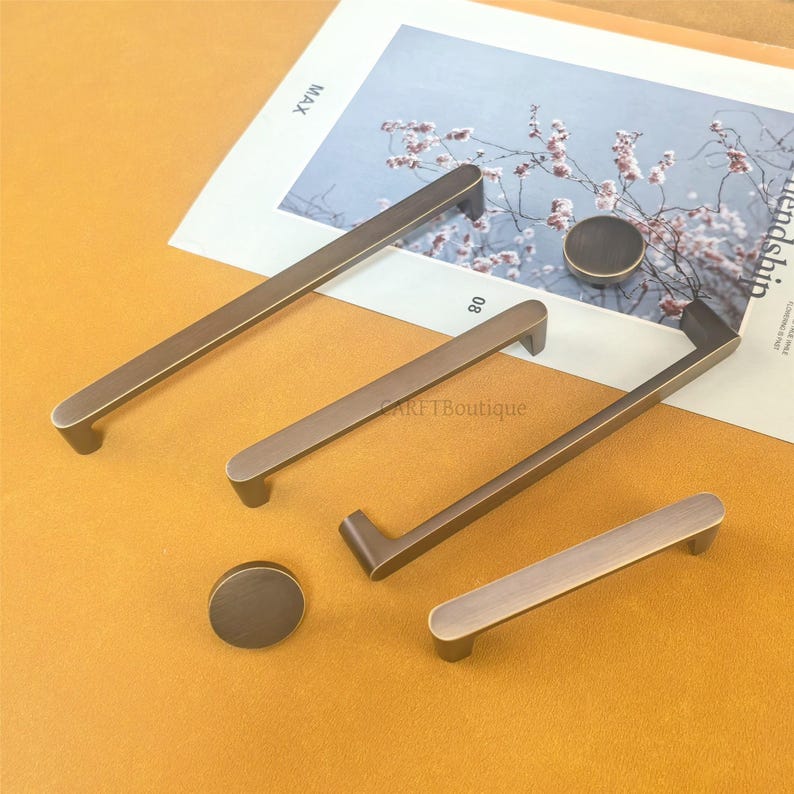 May include: A selection of bronze-coloured cabinet handles and knobs. The set includes different handle designs and circular knobs. The items are displayed on a light surface, with a magazine visible in the background. The handles vary in length.