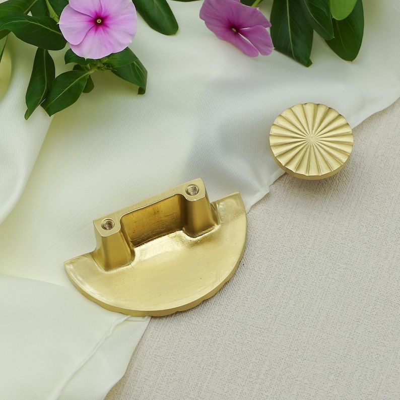 May include: Two brass cabinet hardware pieces. One is a half-moon shape with two mounting posts, and the other is a round knob with a sunburst design. The hardware is displayed on a white cloth with pink flowers and green leaves.