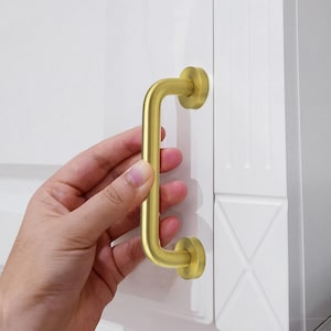 May include: A gold-coloured cabinet pull handle with a curved design. The handle is attached to a white cabinet door. The handle has a cylindrical bar and circular mounting plates. A hand is holding the handle.