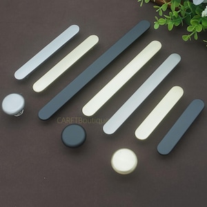 Pode incluir: Uma coleção de puxadores e botões de armário em prata, ouro e preto. A imagem mostra uma variedade de puxadores de barra e botões redondos, adequados para projetos de mobiliário e melhorias domésticas, dispostos sobre uma superfície castanha.