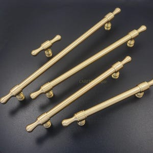 K&ouml;nnte beinhalten: Eine Sammlung von messingfarbenen Schrankgriffen und -kn&ouml;pfen. Die Hardware verf&uuml;gt &uuml;ber eine zylindrische Stange mit dekorativen Endst&uuml;cken und einem zentralen Befestigungspunkt. Die Griffe sind unterschiedlich gro&szlig;, ein kleiner T-f&ouml;rmiger Griff ist enthalten.