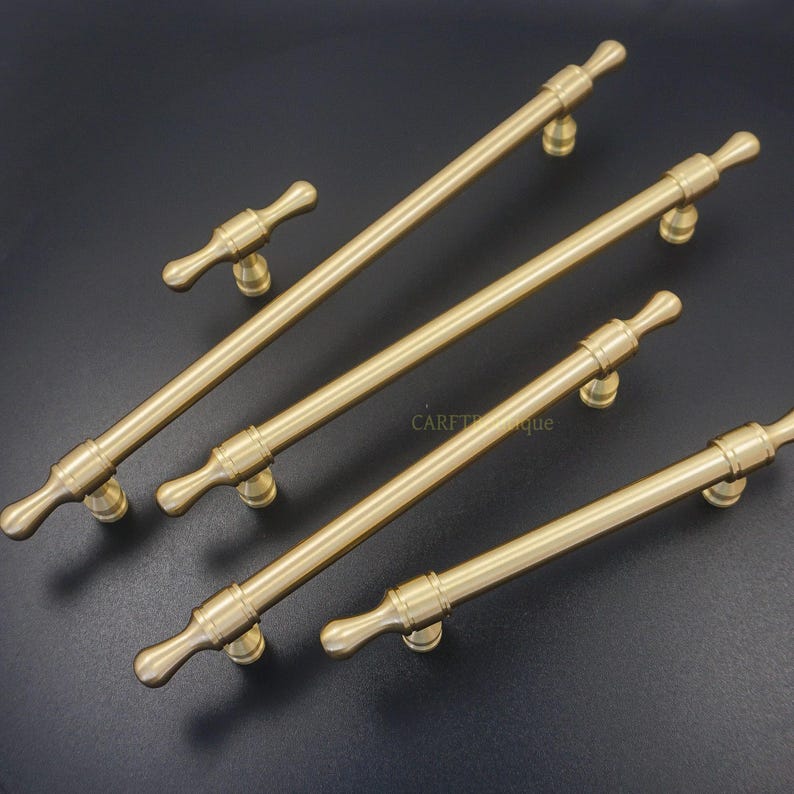 K&ouml;nnte beinhalten: Eine Sammlung von messingfarbenen Schrankgriffen und -kn&ouml;pfen. Die Hardware verf&uuml;gt &uuml;ber eine zylindrische Stange mit dekorativen Endst&uuml;cken und einem zentralen Befestigungspunkt. Die Griffe sind unterschiedlich gro&szlig;, ein kleiner T-f&ouml;rmiger Griff ist enthalten.