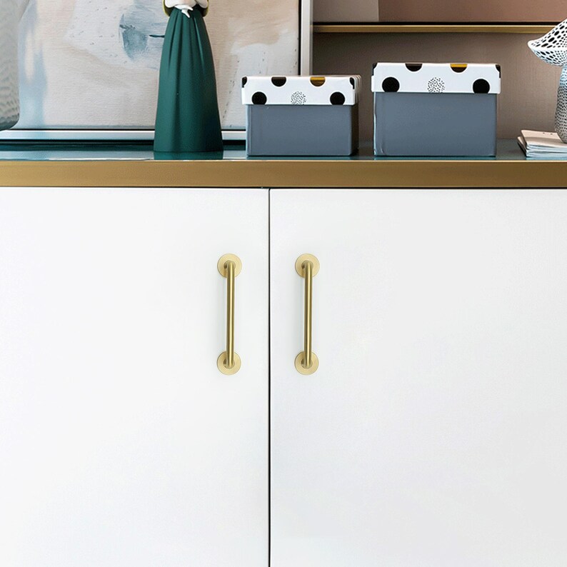 May include: White cabinet doors with two gold-colored bar handles. Above, two gray boxes with black polka dot lids sit on a shelf. A dark green decorative object and a painting are also visible.