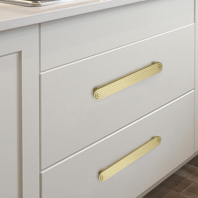 Puede incluir: Primer plano de un mueble de cocina con dos cajones. Cada caj&oacute;n tiene un tirador horizontal de color dorado con un dise&ntilde;o acanalado. Las puertas del armario son de color gris claro y los tiradores est&aacute;n centrados en cada caj&oacute;n.