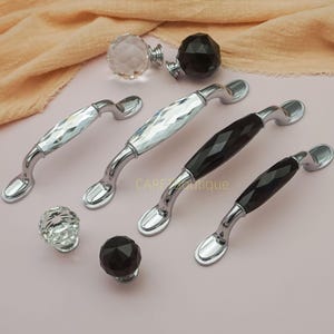 Könnte beinhalten: Eine Sammlung von Schrankbeschlägen, darunter Kristall- und schwarze facettierte Knöpfe und Griffe. Die Griffe haben ein geschwungenes Design mit silbernem Finish. Die Knöpfe und Griffe werden auf einer hellrosa Oberfläche präsentiert.