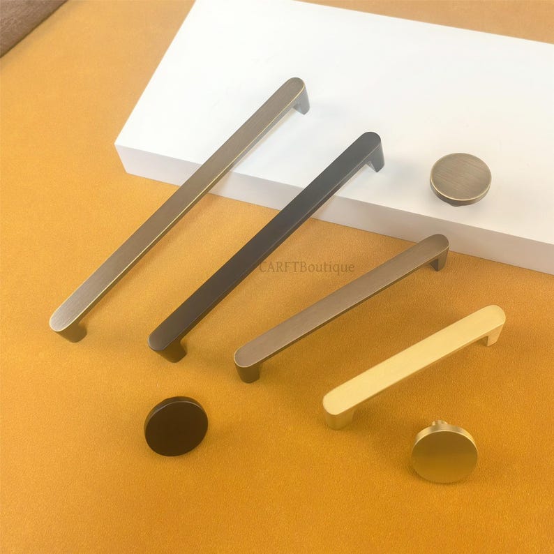 May include: A selection of contemporary cabinet handles and knobs in metallic finishes. Features long bar pulls in bronze, black, and gold, plus matching round knobs. The hardware is presented on a white surface against a yellow background.