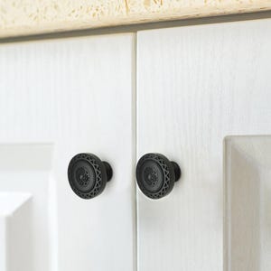May include: Two black cabinet knobs with a decorative floral and geometric pattern. The knobs are round and mounted on white cabinet doors. The knobs have a matte finish and are made of metal.