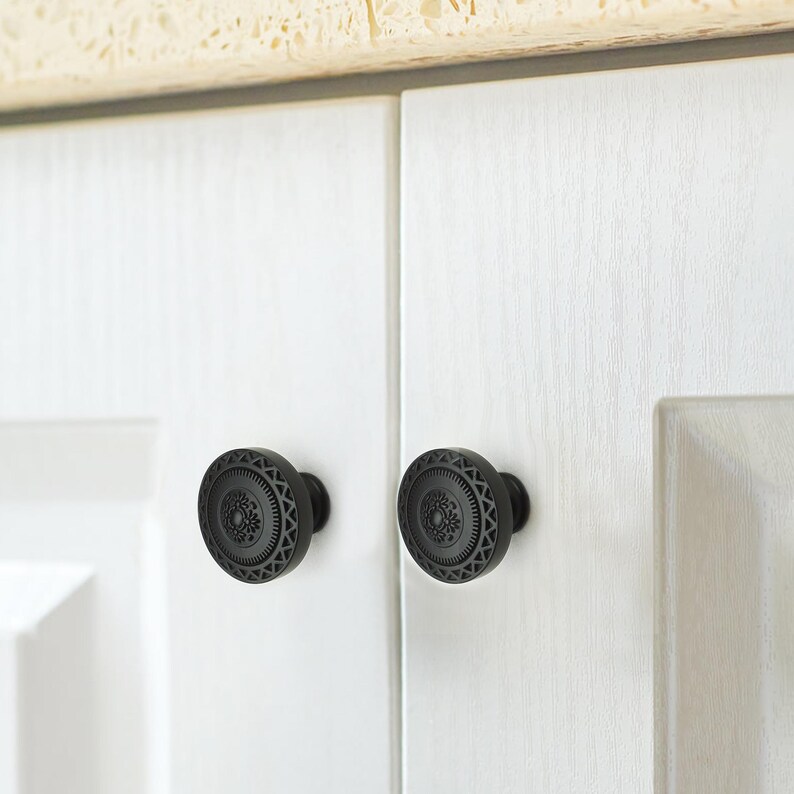 May include: Two black cabinet knobs with a decorative floral and geometric pattern. The knobs are round and mounted on white cabinet doors. The knobs have a matte finish and are made of metal.