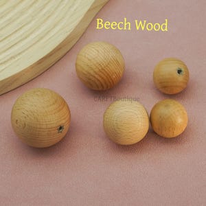 May include: A collection of beech wood spheres in various sizes. The spheres have a natural wood grain pattern and are light brown in color. Some spheres have a small hole drilled through them. The text "Beech Wood" is visible in the image.