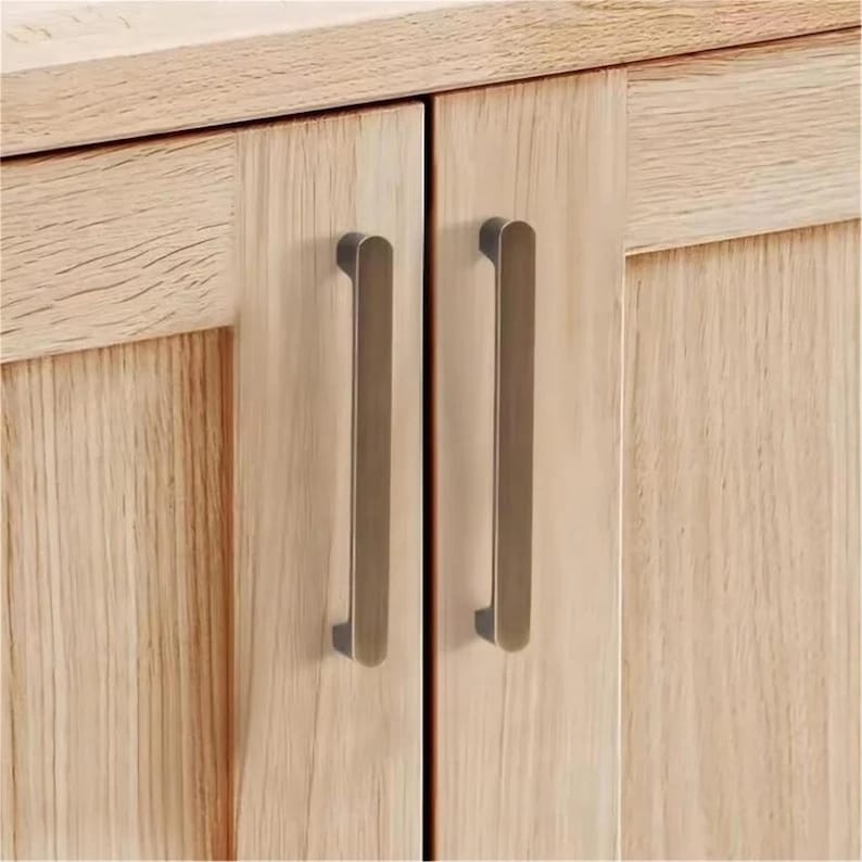 May include: Close-up of a light-coloured wooden cabinet with two bronze-coloured, rectangular bar handles. The cabinet doors have a vertical wood grain pattern. The handles are mounted vertically on the doors.