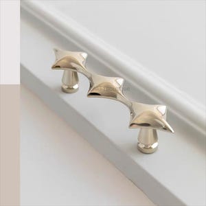 以下が含まれることがあります： ユニークな幾何学模様の、磨かれたシルバーカラーのキャビネットプル。プルは、湾曲したバーで接続された3つのダイヤモンド型の要素を備えています。「ARTfBoutique」の文字がプルに表示されます。このハードウェアは引き出しやキャビネットに適しています。