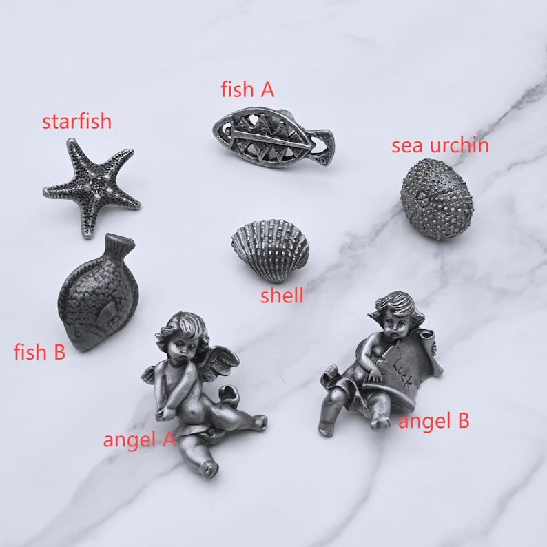 Peut inclure: Une collection de petits objets d&eacute;coratifs argent&eacute;s. Les articles comprennent une &eacute;toile de mer, deux poissons, un coquillage, un oursin et deux figurines d'anges. Les figurines d'anges tiennent des parchemins. Les articles sont dispos&eacute;s sur une surface en marbre blanc.