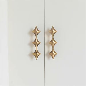 以下が含まれることがあります： 白いキャビネットドアに取り付けられた、星型のデザインの真鍮製キャビネットプルが2つあります。プルは、垂直に積み重ねられた一連のダイヤモンドの形をしています。キャビネットドアは細い黒い線で区切られています。