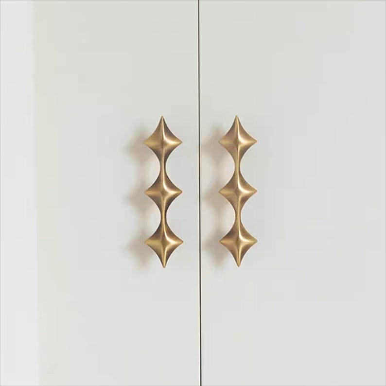 以下が含まれることがあります： 白いキャビネットドアに取り付けられた、星型のデザインの真鍮製キャビネットプルが2つあります。プルは、垂直に積み重ねられた一連のダイヤモンドの形をしています。キャビネットドアは細い黒い線で区切られています。